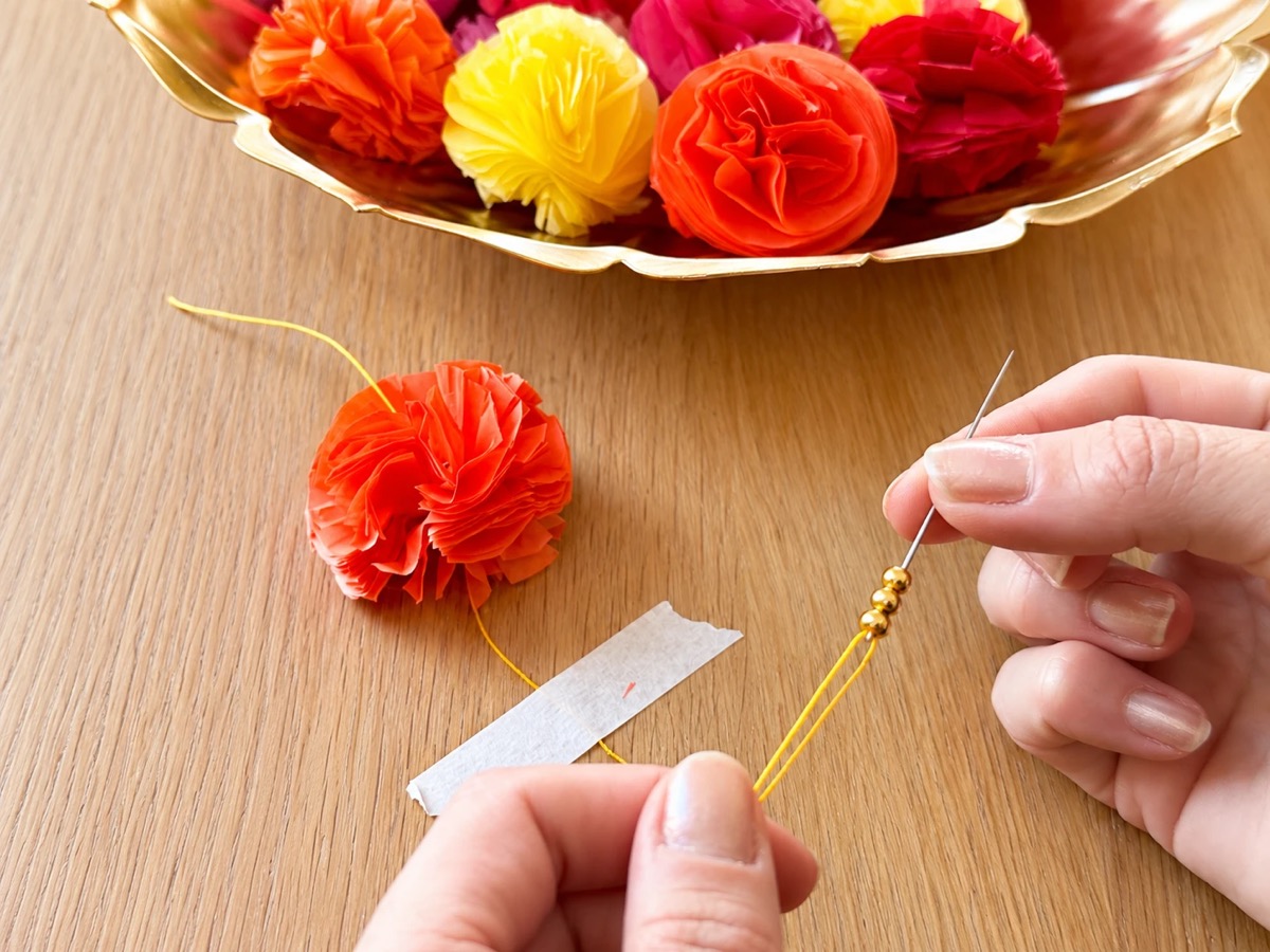 Sliding three gold beads onto the needle above an orange tissue paper flower, with a bowl of flowers behind.