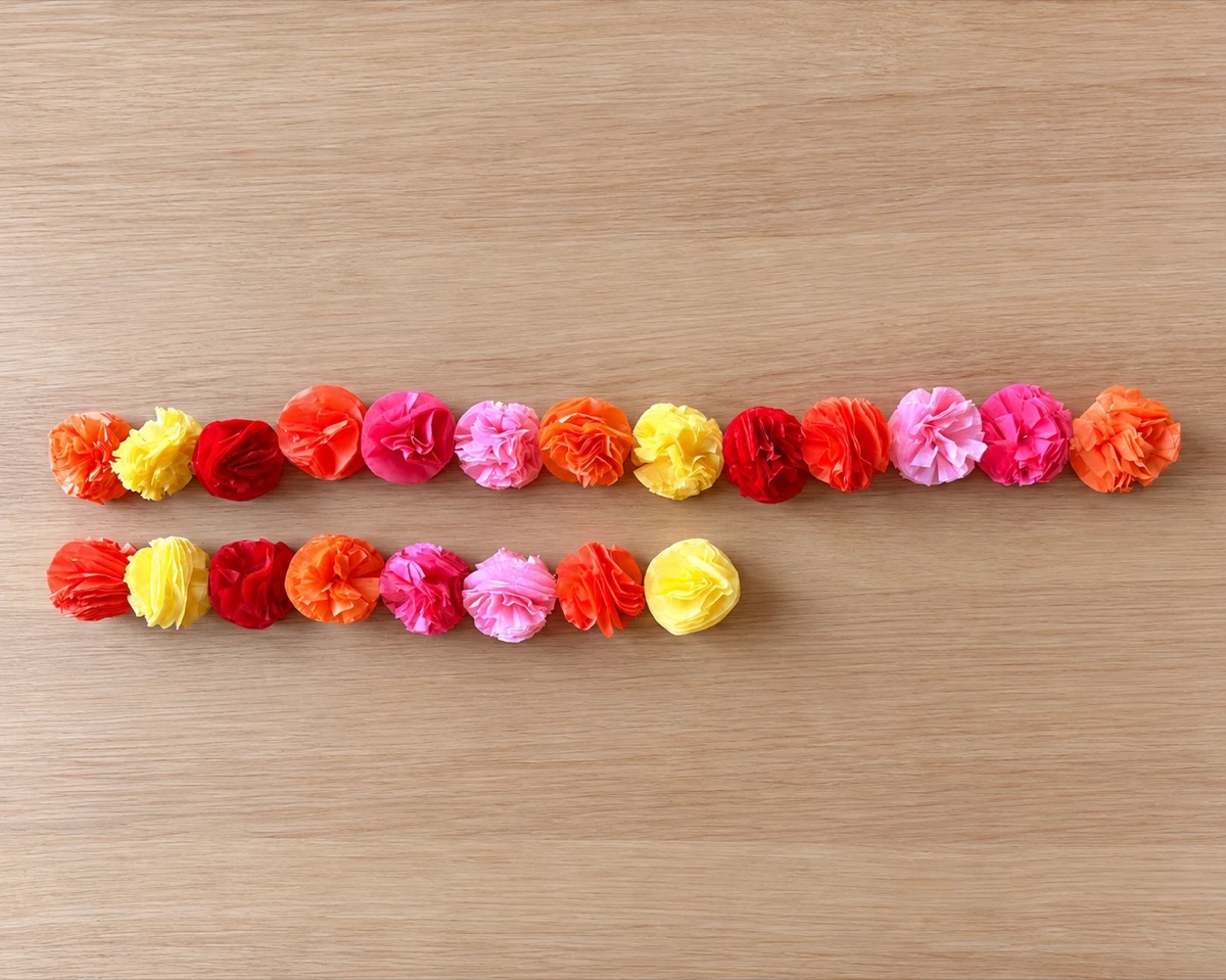 Tissue paper marigolds laid out in two rows in color order: 13 for the long garland and 8 for the short one.