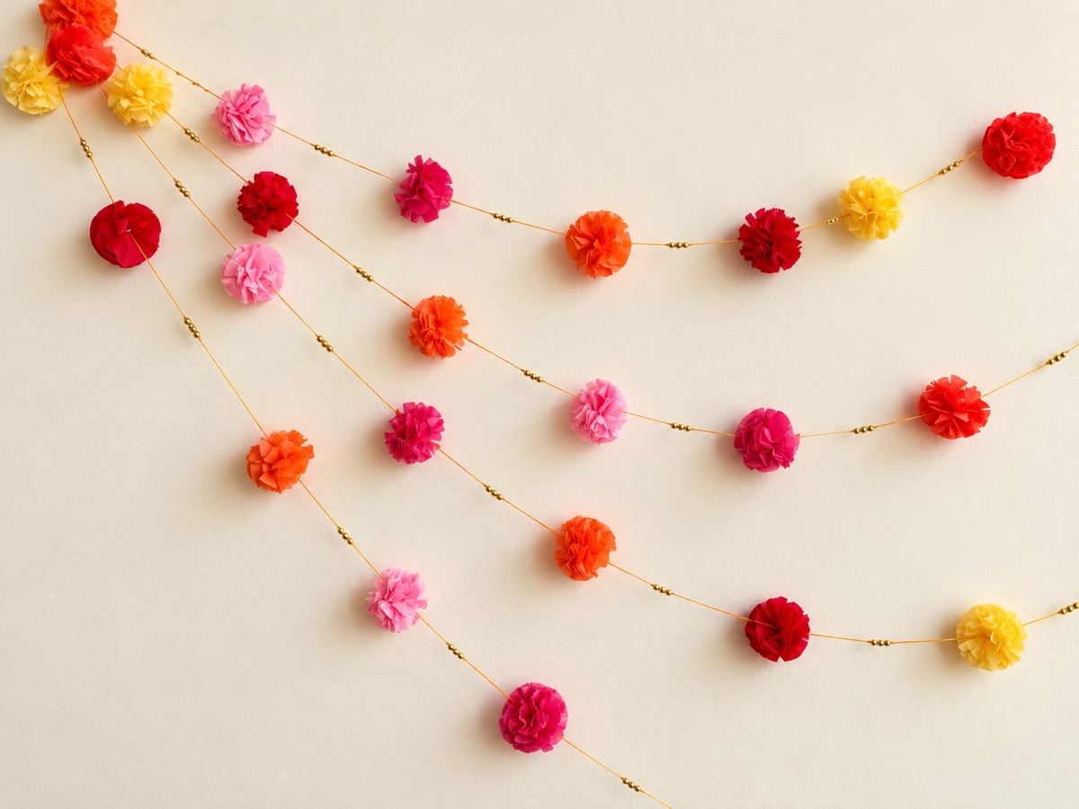 Two tissue paper marigold garlands in pink, orange and yellow with gold beads, hung on a cream wall.
