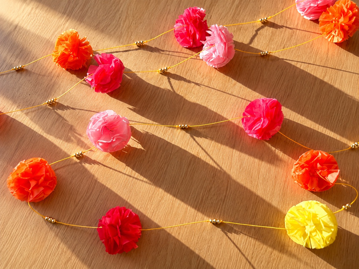 Two tissue paper marigold garlands laid out in sunlight, pink, orange, red and yellow flowers with gold beads on orange cord