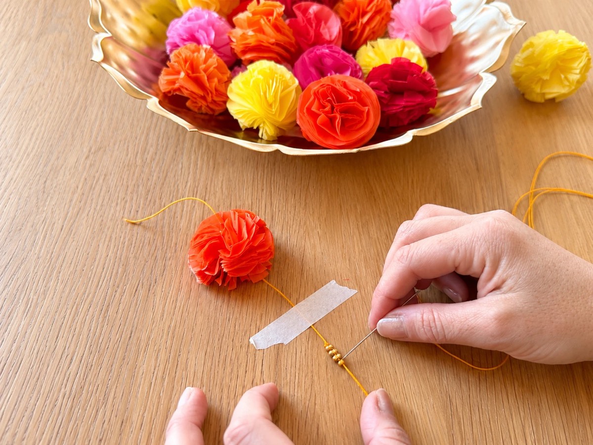 Tying a triple knot just above three gold beads on the orange cord, with the cord taped to the table and a bowl of flowers behind.