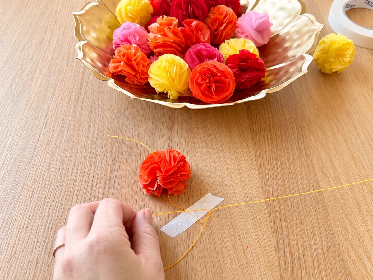 Tying a knot in the orange cord above an orange tissue paper flower, with the cord taped to the table and a bowl of flowers behind.