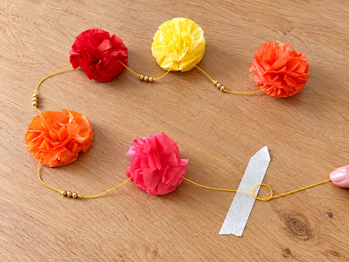 A partly strung garland with five flowers, gold beads between them, and the cord taped to the table.