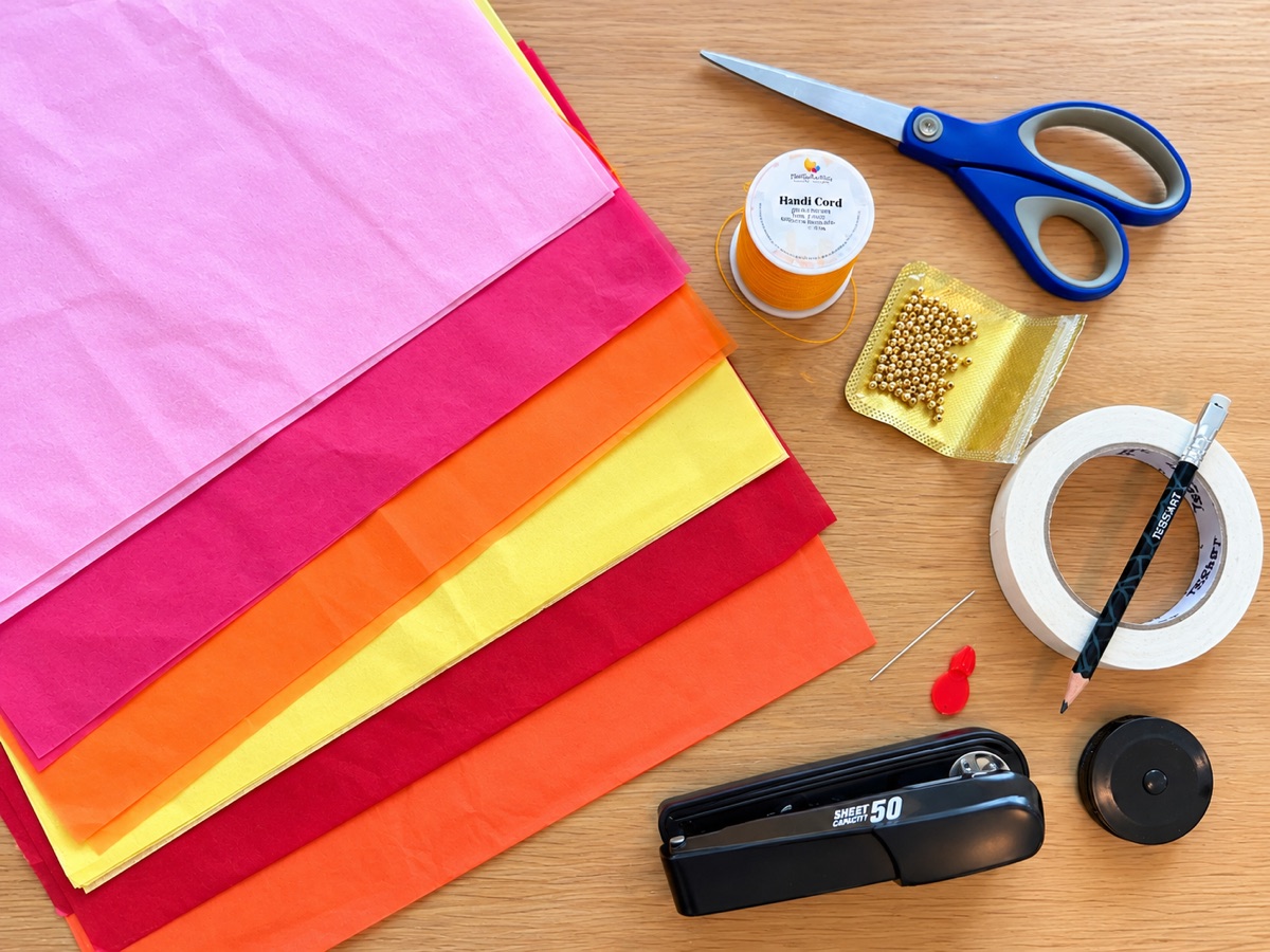 Everything you need laid out on a wooden table: tissue paper in six colors, scissors, orange waxed cord, gold beads, masking tape, a pencil, a needle and threader, a stapler, and a tape measure.