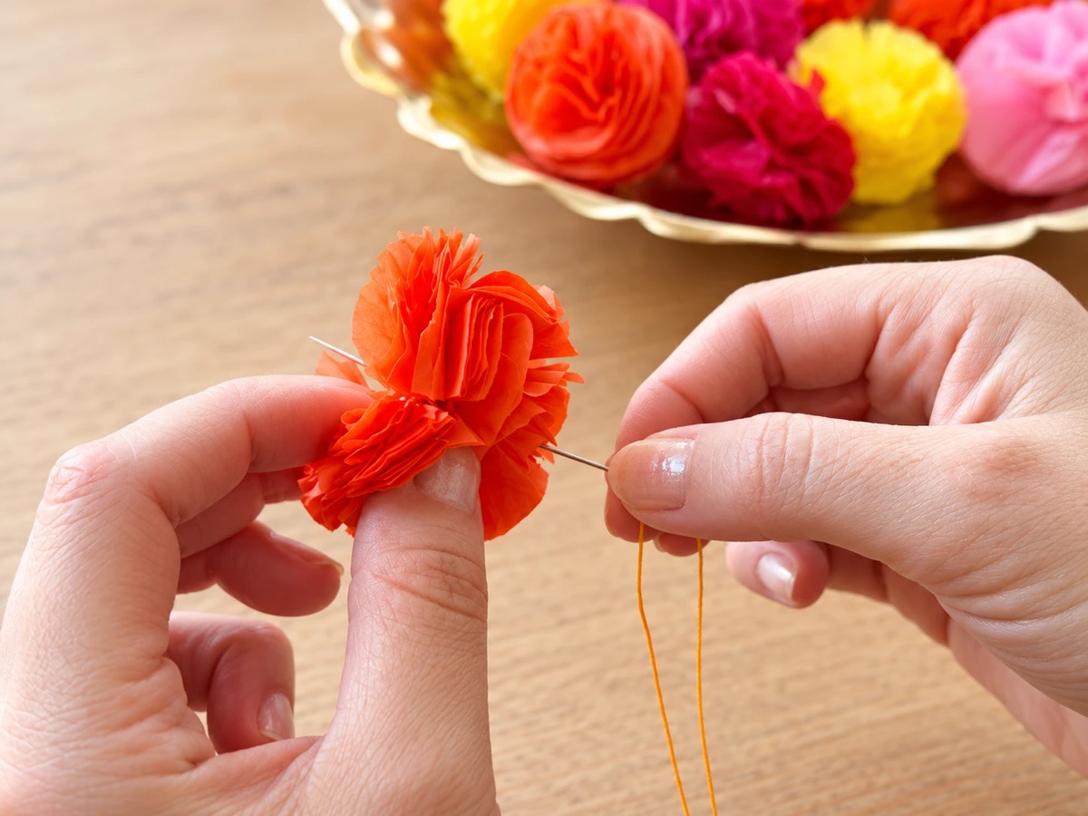 Pushing a threaded needle through the center of an orange tissue paper flower, with a tray of finished flowers behind.